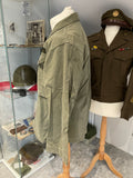 Green military-style pants on a mannequin with a brown jacket and helmet in the background.